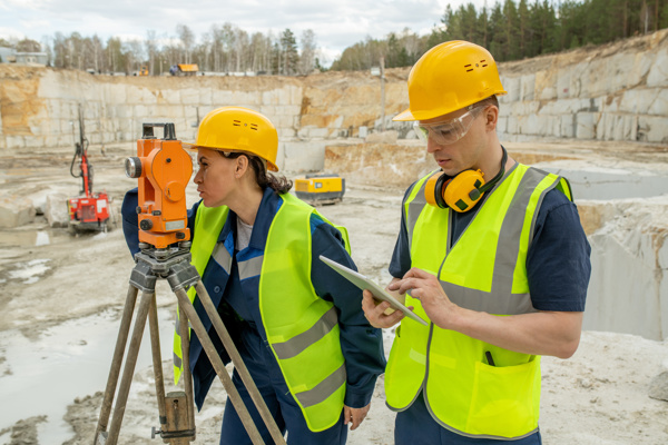 Person with a hard hat is looking through a geodetic station, another person is looking at his tablet