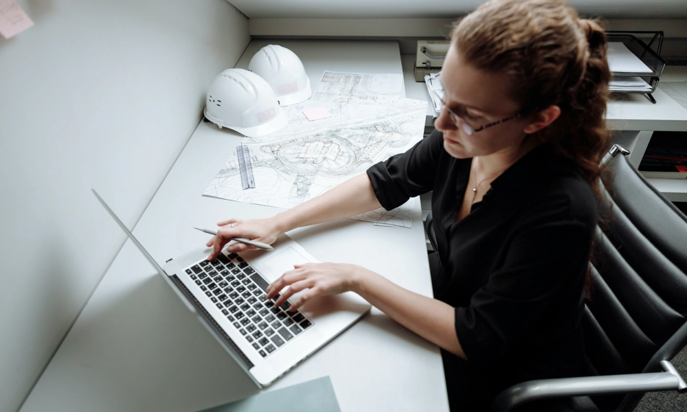Engineer working at her laptop with building drawings and hard hats on the table next to her