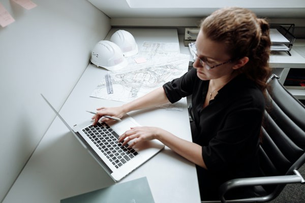Engineer working at her laptop with building drawings and hard hats on the table next to her