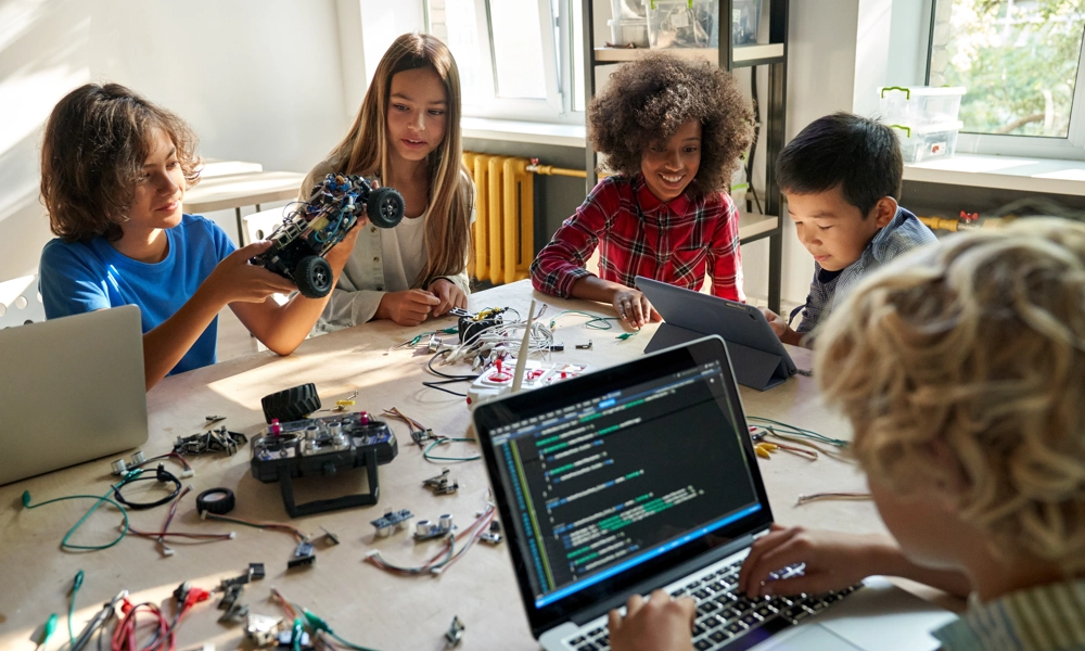 Five children sitting around a table are working on laptops, tablets and circuits for STEM toys
