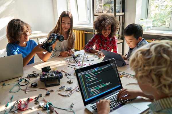 Five children sitting around a table are working on laptops, tablets and circuits for STEM toys