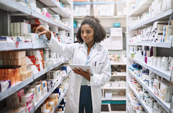 Person with a white coat is holding a notepad and looking at medications on the shelves