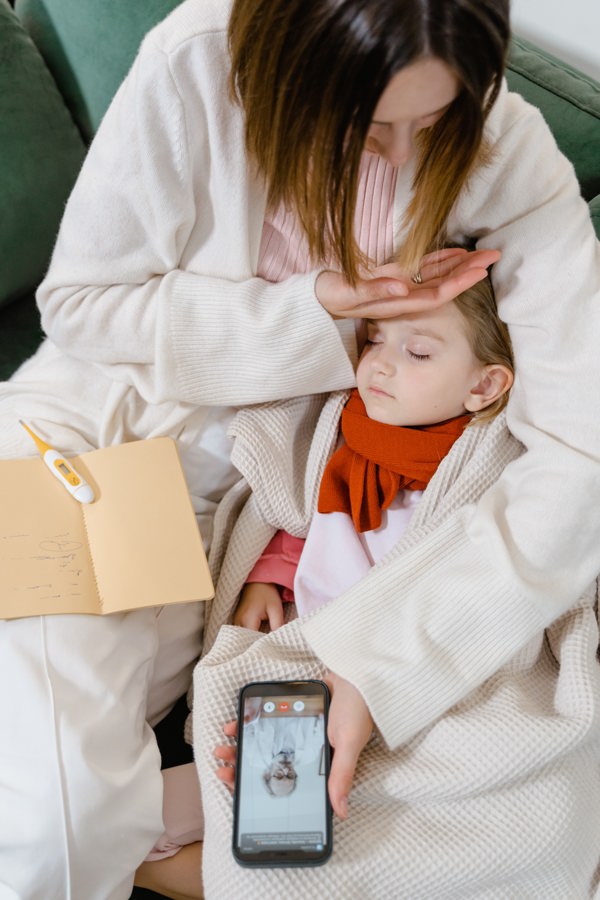 Woman has her hand on the sleeping child's forehead while her mobile phone shows a doctor on the screen