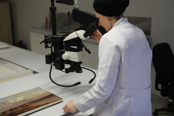 Person in a lab coat looking through the lens of a digital microscope into a painting