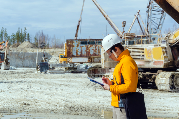 Person with a hard hat taking notes in a construction site with an excavator in the background