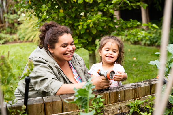 Woman and girl laughing while spraying water to plants in a garden bed