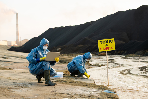 Two people in biohazard suits taking samples from a river, looking at a laptop with a smoking industrial building in the background