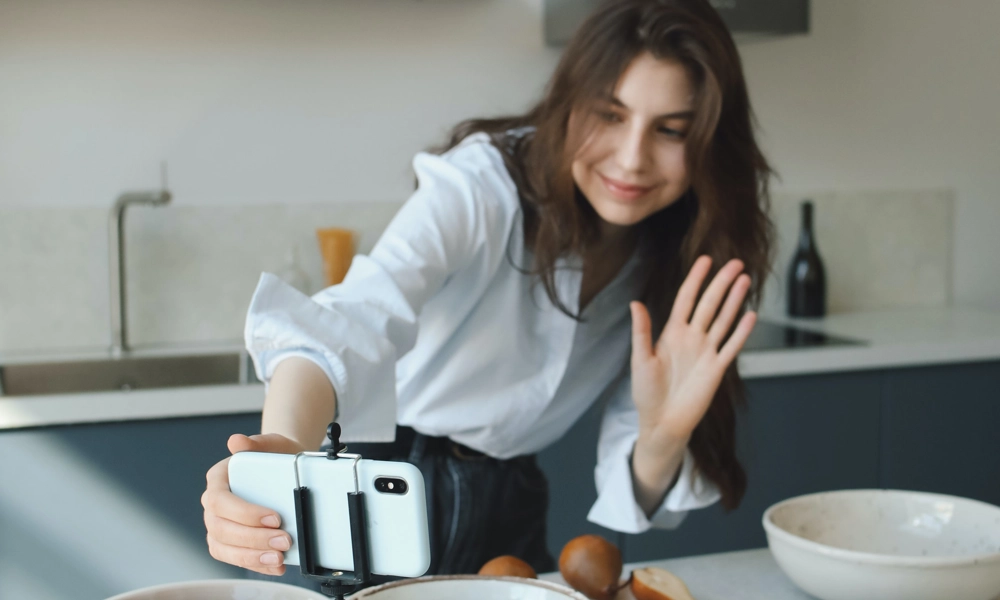 Person waving at her mobile phone set-up to show her and her kitchen